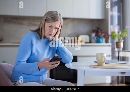 Donna anziana che stringe la testa mentre legge notizie sconvolgenti su uno smartphone, mostrando preoccupazione, stress e tristezza in un ambiente tranquillo in cucina Foto Stock