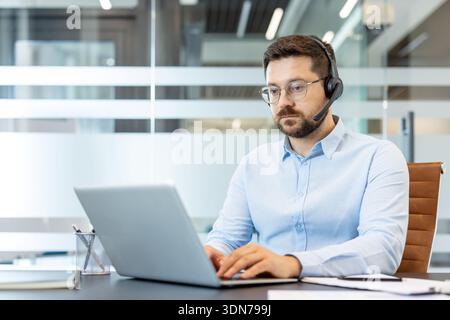 Uomo professionista in camicia con bottoni che indossa una cuffia con microfono, lavora su un computer portatile in un ufficio moderno e fornisce assistenza tecnica remota e assistenza clienti virtuale durante le videochiamate Foto Stock