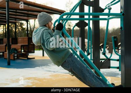 Una donna determinata esegue un pull-up supportato su una palestra esterna. La prospettiva laterale mostra tutta la tensione e il coinvolgimento del corpo, evidenziando la dedizione a rafforzare la schiena e le braccia in condizioni climatiche fredde Foto Stock