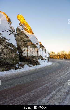 Una strada si snoda intorno a una grande formazione rocciosa in inverno. La neve copre il terreno e la luce del sole proietta un caldo bagliore sulla roccia. Gli alberi fiancheggiano lo sfondo sotto un cielo limpido. Foto Stock