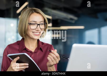 Giovane donna d'affari che indossa gli occhiali sorridenti e tiene in mano un notebook e una penna durante una videoconferenza online o una riunione su un notebook in un ambiente di lavoro moderno Foto Stock