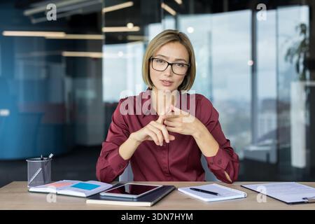 Donna d'affari sicura che indossa occhiali da vista e una camicia marrone seduta alla scrivania, gestando con le mani durante una riunione virtuale in un moderno ufficio open space Foto Stock