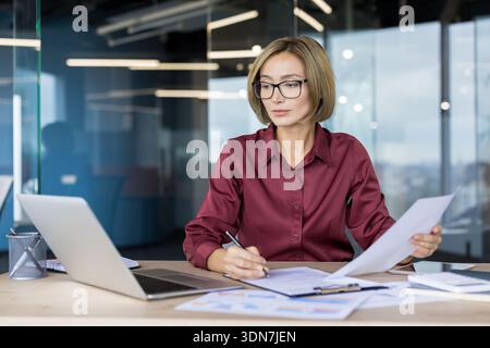 Donna d'affari fiduciosa che lavora diligentemente, organizzando i documenti e firmando accordi alla sua scrivania in uno spazio di lavoro contemporaneo, dimostrando attenzione e competenza nel suo ruolo professionale Foto Stock