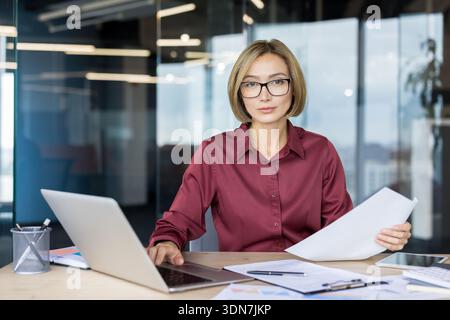 Donna d'affari sicura che indossa occhiali da vista e una camicia marrone, guarda la fotocamera mentre lavora su un laptop e tiene in mano documenti, concentrandosi sulla gestione e sul successo aziendale nel suo ambiente di lavoro moderno Foto Stock