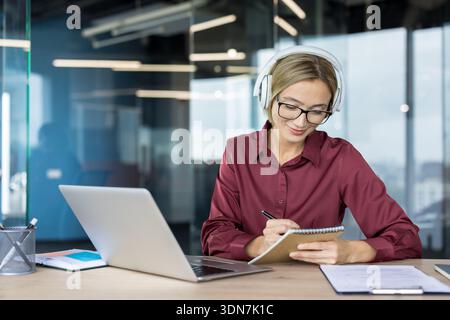 Giovane donna che indossa cuffie e occhiali, guarda un webinar sul laptop, scrive appunti su un notebook, studia o lavora in remoto in un ufficio moderno Foto Stock
