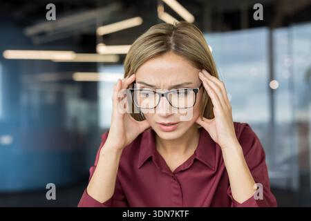 Giovane donna che soffre di mal di testa e stress, che tiene le tempie, soffre di disagio e dolore mentre gestisce una giornata lavorativa impegnativa in un ufficio moderno Foto Stock