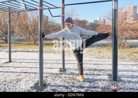 L'uomo asiatico anziano con cappuccio e berretto si allunga su un palo metallico al campo di allenamento in strada nel parco cittadino nella soleggiata giornata invernale. Foto Stock