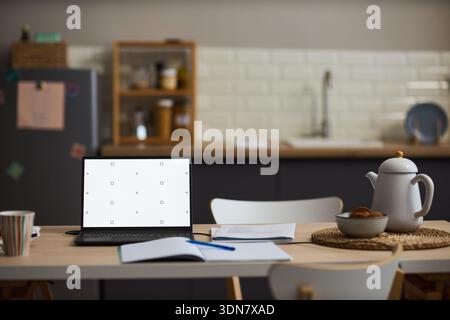 Laptop aperto con schermo vuoto in piedi sul tavolo della cucina accanto a notebook aperto, penna, documenti, teiera e tazza, moderno sfondo della cucina domestica visibile con una messa a fuoco morbida Foto Stock