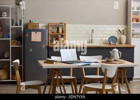 Moderno interno della cucina con tavolo da pranzo in legno con laptop aperto, notebook, tazza, teiera e piatto, sedie vuote intorno al tavolo, scaffali e frigorifero sullo sfondo Foto Stock