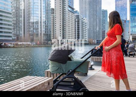 Una madre incinta va a fare una passeggiata lungo il fiume in una città urbana con una seggiolone per bambini in una giornata di sole Foto Stock