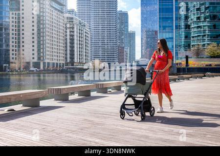 Una madre incinta va a fare una passeggiata lungo il fiume in una città urbana con un passeggino in una giornata di sole Foto Stock