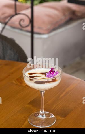 panna cotta alla vaniglia setosa e cremosa servita in un elegante calice di vetro, splendidamente condita con un ricco goccio di cioccolato e un delicato fl commestibile viola Foto Stock