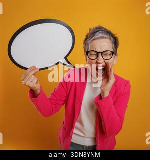 Una donna entusiasta con i capelli grigi corti grigia grida gioiosamente mentre tiene una bolla bianca su uno sfondo giallo brillante, trasmettendo eccitazione Foto Stock