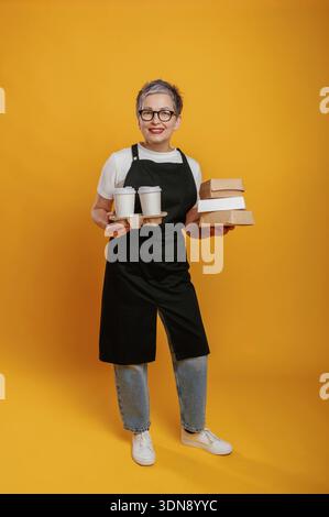 Una donna allegra che indossa grembiule e bicchieri neri tiene in mano contenitori da asporto e tazze su uno sfondo giallo brillante, che rappresenta il cibo servito Foto Stock