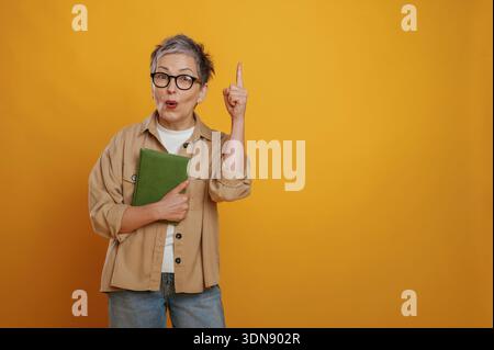 Una donna anziana con capelli e occhiali grigi tiene in mano un notebook verde mentre punta verso l'alto, trasmettendo entusiasmo e ispirazione contro un giallo luminoso Foto Stock