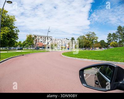 Le Touquet, Francia - 22 agosto 2023: POV dal veicolo che mostra lo specchio retrovisore su una passeggiata pavimentata rosa curvilinea con erba, lampioni, alberi e. Foto Stock