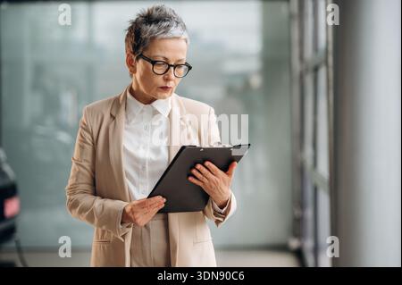 Una donna matura con un abito di colore chiaro è incentrata sulla revisione dei documenti su una clipboard mentre si trova in un elegante spazio ufficio con grandi finestre e. Foto Stock