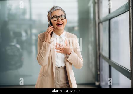 Una donna anziana vestita con un abito alla moda ride gioiosamente mentre parla sul suo smartphone, circondata da un luminoso spazio ufficio con un grande vento Foto Stock