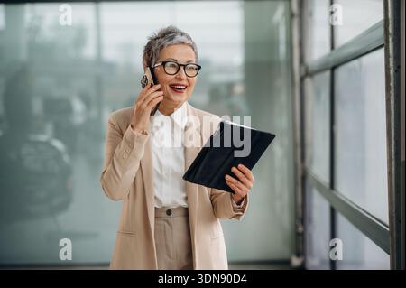 Una donna matura con i capelli grigi corti sorride mentre è impegnata in una conversazione telefonica e tiene in mano un tablet in un moderno ufficio con grandi dimensioni Foto Stock