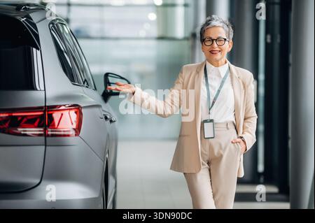 Una donna anziana vestita con un elegante abbigliamento da lavoro si erge accanto a un veicolo moderno in uno showroom luminoso, presentando con sicurezza l'auto e dimostrando Foto Stock