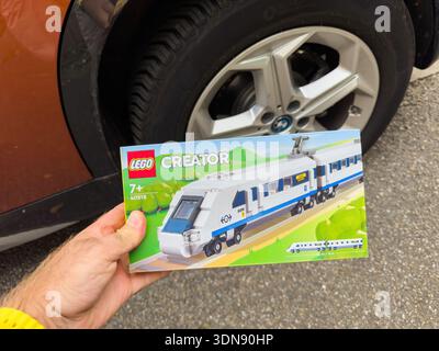 Parigi, Francia - 17 settembre 2025: Scatola giocattolo LEGO Creator con aggancio a mano e modello di treno passeggeri bianco e blu davanti alla ruota arancione del veicolo BMW e allo pneumatico sulla superficie dell'asfalto. Foto Stock