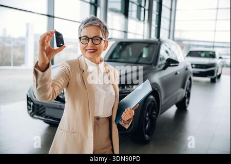 Una donna anziana vestita con un abito alla moda sorride mentre tiene in mano le chiavi dell'auto in uno showroom automobilistico contemporaneo, circondato da veicoli di lusso, hig Foto Stock