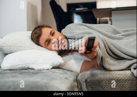 L'uomo con i capelli marrone chiaro sta sdraiandosi su un divano, avvolto in una morbida coperta, tenendo in mano un telecomando, e si gode un momento di relax guardando la televisione Foto Stock