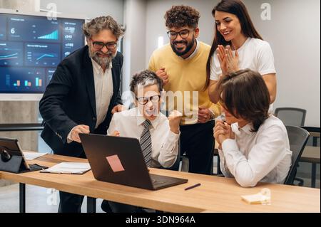 Gruppo eterogeneo di professionisti che festeggiano con gioia un progetto di successo in un ufficio moderno, riuniti attorno a un notebook che mostra grafici e g Foto Stock