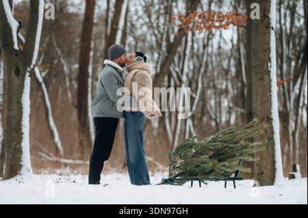 Coppia afroamericana che condivide un bacio amorevole in una foresta innevata, circondata da alberi alti, con un albero di Natale appena tagliato su una slitta, che incarna il Foto Stock