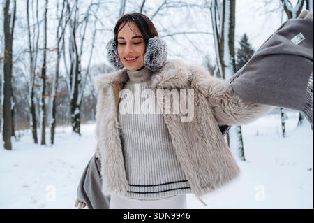 Modello femminile vestito con comodi abiti invernali, sorridente in una foresta innevata, con eleganti cuffie e cappotto in pelliccia sintetica, che catturano l'essenza del vento Foto Stock