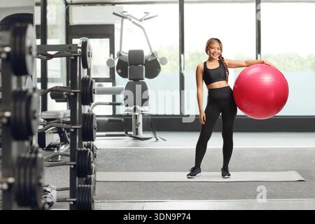 Giovane donna asiatica in costume fitness che posa con una palla rossa in palestra Foto Stock