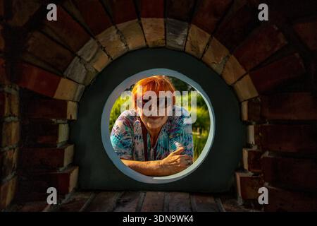 Una donna felice guarda attraverso una finestra rotonda a Ogrody Kapias, Polonia, godendo di fiori colorati, vegetazione lussureggiante e una gioiosa tranquillo giardino. Foto Stock