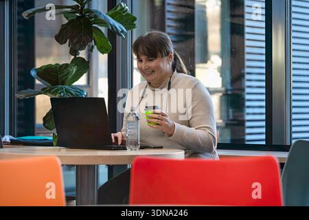 Una donna professionista con i capelli castani lavora al tavolo, utilizza un computer portatile e sorseggia un drink, circondato da uno spazio di lavoro vivace e moderno Foto Stock