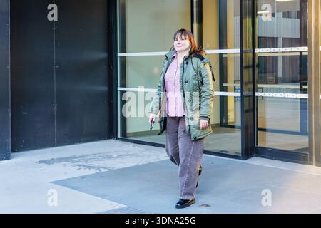 Una donna con giacca verde e pantaloni grigi, cammina fuori da una struttura contemporanea, incarnando lo stile urbano e la fiducia in un ambiente vivace Foto Stock