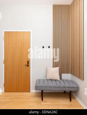 Un ingresso moderno caratterizzato da una porta in legno naturale, una panchina grigia testurizzata con un cuscino beige e una parete verticale con accenti in legno. Foto Stock