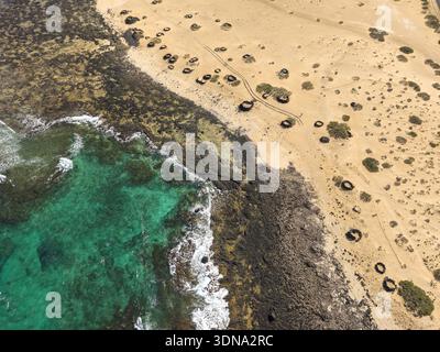 Vista aerea delle acque turchesi che incontrano la costa rocciosa e aspra e la spiaggia sabbiosa punteggiata da formazioni scure e circolari, il parco nazionale di Corralejo e Fue Foto Stock