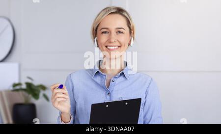 Donna sorride mentre tiene in mano una clipboard e una penna in un ambiente Office moderno Foto Stock