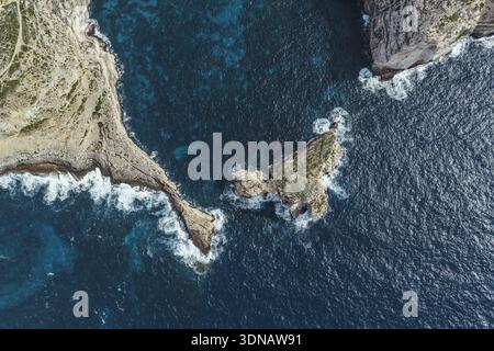 Vista aerea della costa frastagliata, dove le scogliere incontrano il mare blu profondo, le onde che si infrangono contro le coste rocciose, la baia di Dwejra, Malta. Foto Stock
