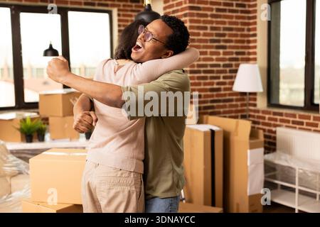 Fidanzata caucasica e fidanzato afroamericano che festeggia con abbraccio in un nuovo appartamento. Coppia felice e variegata che abbraccia gioiosamente dopo aver completato l'acquisto di una moderna proprietà residenziale. Foto Stock