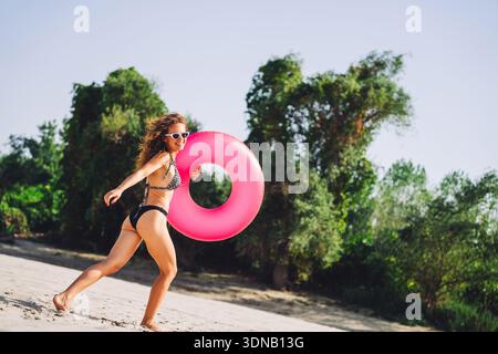Bella giovane donna che si diverte sulla spiaggia sabbiosa con galleggianti rosa in estate Foto Stock