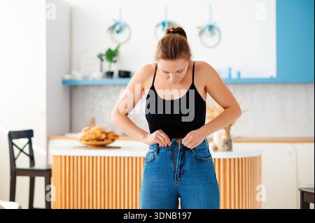 Una giovane donna con una canotta nera lotta per chiudere i jeans blu in un ambiente luminoso da cucina Foto Stock