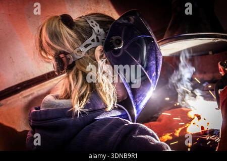 Saldatore industriale che esegue la saldatura ad arco su metallo in un ambiente di officina con scintille di saldatura brillanti, fumo e casco protettivo. Foto Stock