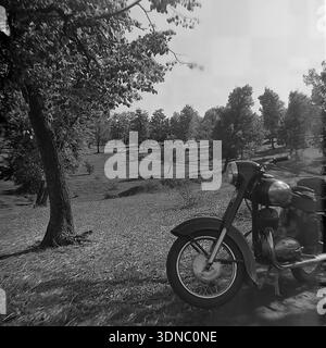 Regione di Donetsk, Ucraina, URSS. 1970 anni Fotografia d'archivio in bianco e nero che raffigura una classica moto d'epoca parcheggiata su una collina. Il primo piano presenta la parte anteriore dettagliata della moto sovietica, mentre lo sfondo rivela un vasto paesaggio ondulato con tortuose strade sterrate, boschetti e un grande albero frondoso che getta ombre sull'erba. Questa scena nostalgica cattura l'essenza del viaggio rurale e del tempo libero all'aperto nella tranquilla regione del Donbass. L'immagine riflette un senso di libertà e tranquillità durante l'era sovietica. Foto Stock