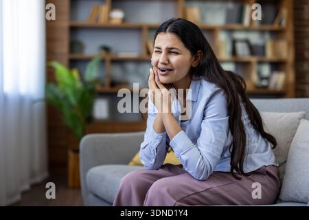 Giovane donna su un divano che tiene la guancia nel dolore, soffre di un forte mal di denti a casa e ha bisogno di cure dentistiche urgenti e sollievo Foto Stock