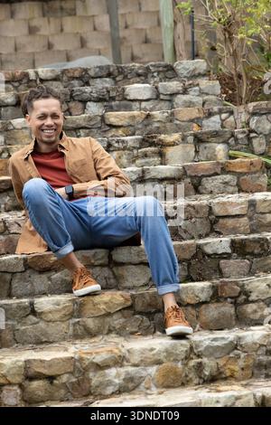 Uomo seduto e appoggiato su gradini di pietra sulla terrazza all'aperto con smartwatch e jeans Foto Stock