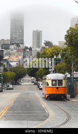 Una storica funivia di San Francisco si sposta lungo una ripida strada residenziale costeggiata da alberi e condomini in una nebbiosa giornata a San Francisco Foto Stock