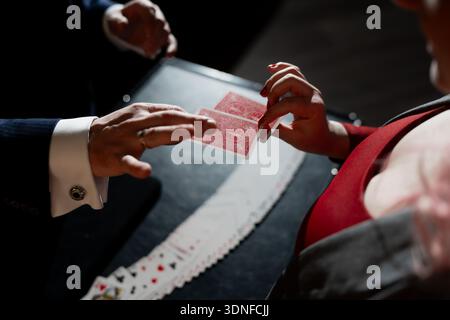 Mago che esegue un trucco di carte per una donna in abito rosso, scambia le mani della carta da gioco sopra il tavolo con il mazzo ventilato, luci e ombre drammatiche Foto Stock