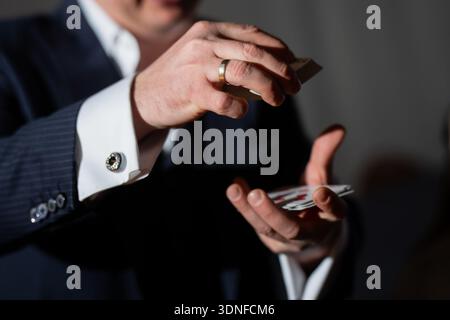 Primo piano del mago delle carte che si occupa e manipola le carte da gioco con le mani, gemelli in tuta e anello d'oro visibili sotto i riflettori Foto Stock