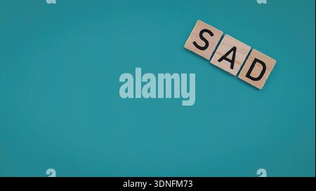 Un'immagine semplice con tre piastrelle di legno disposte per scrivere "SAD" su uno sfondo blu pieno. Foto Stock