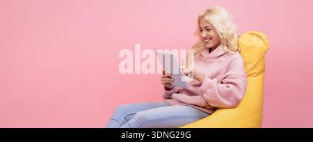 Una giovane donna ama il tablet seduto su una borsa con fagioli gialli nella stanza rosa Foto Stock